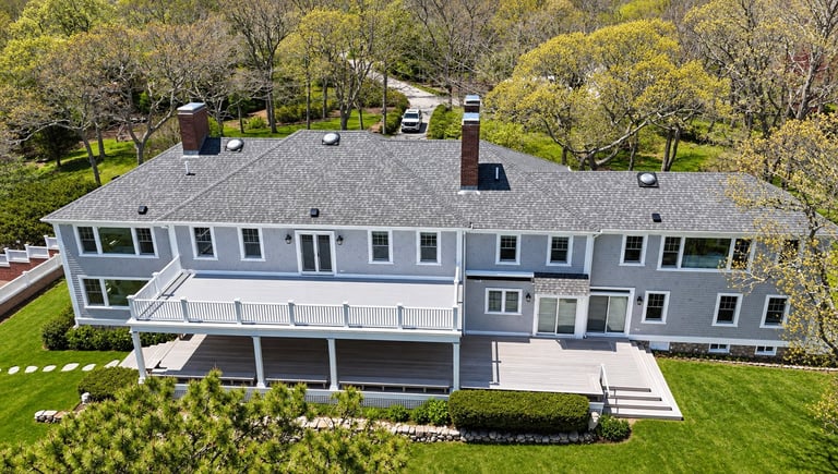 Aerial view of a modern gray two-story house with white railings, deck, and landscaped lawn surrounded by trees.