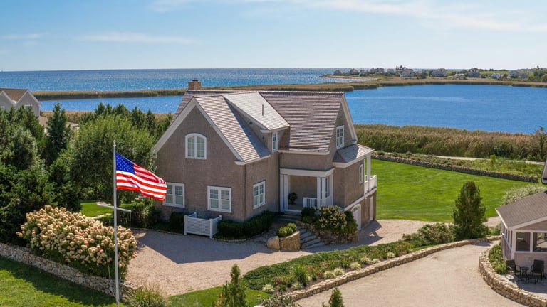Waterfront brick home with American flag on manicured lawn overlooking blue bay water