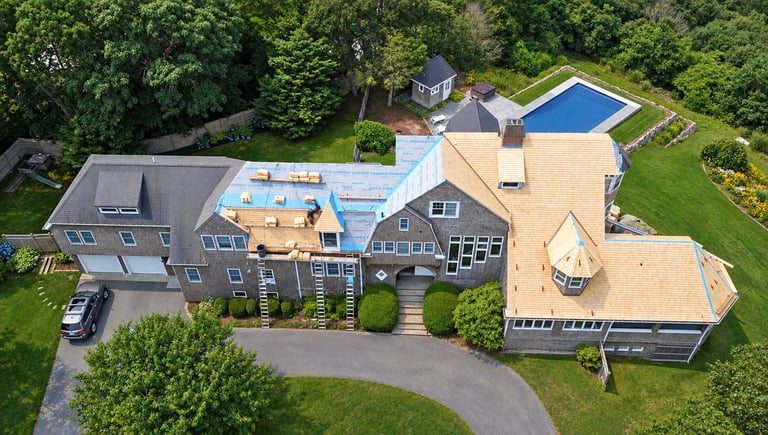 Aerial view of a large residential home with brown shingles, curved driveway, pool, and manicured grounds surrounded by trees.