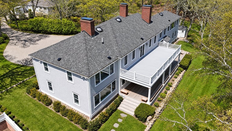 Aerial view of modern white and gray house with deck and manicured lawn surrounded by trees.