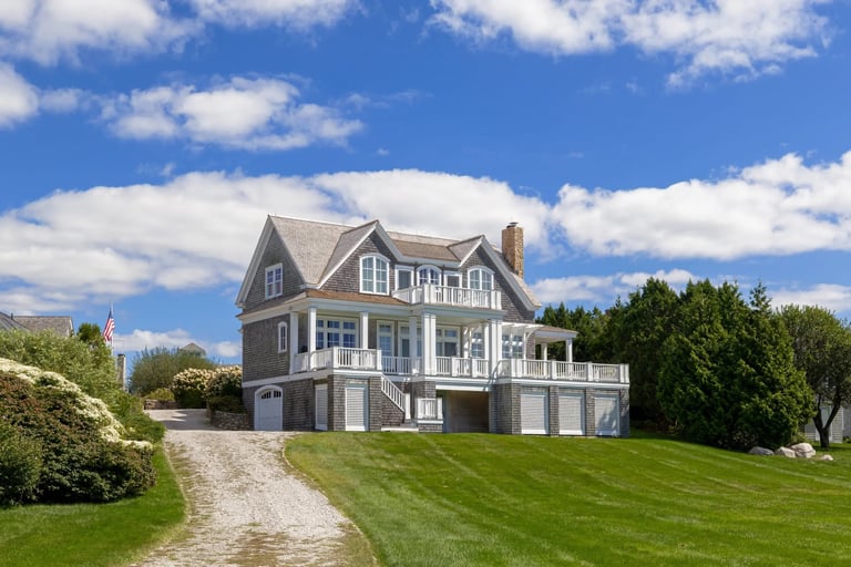 Large gray and blue two-story house with gabled roof and deck, set on manicured lawn with gravel driveway under blue sky