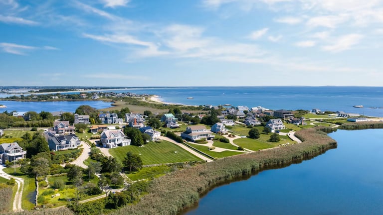 Aerial view of a coastal residential neighborhood with houses, green lawns, and marsh along a blue waterway under clear sky