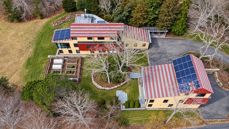 Aerial view of eco-friendly homestead with solar panels, garden beds, and shipping container structures surrounded by trees.