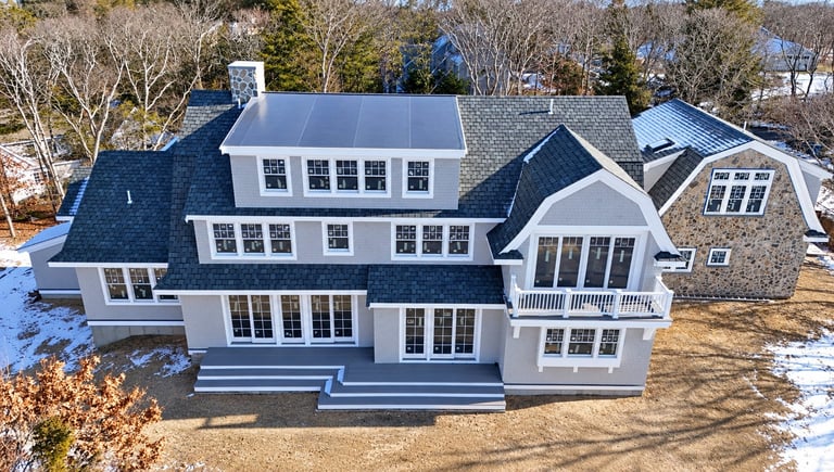 Aerial view of a large white modern house with dark roof, multiple gables, and winter landscaping.