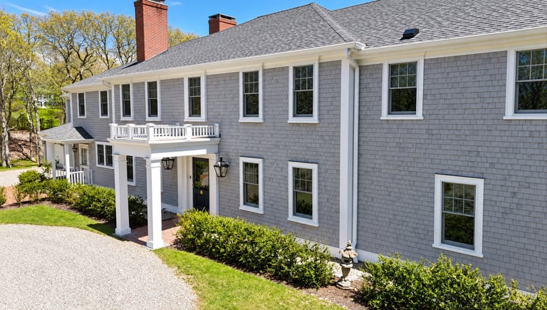 Two-story gray house with white trim, columned porch, and brick chimneys on a landscaped property.