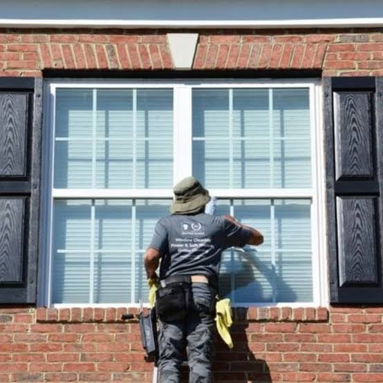 Worker in black shirt and hat cleaning large brick building window with blue shutters on either side