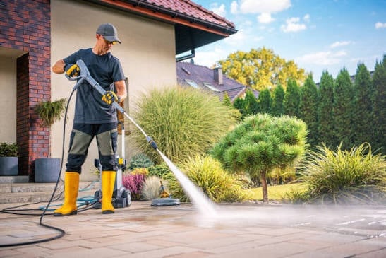 Man in yellow boots and protective gear pressure washing a residential patio with a hose