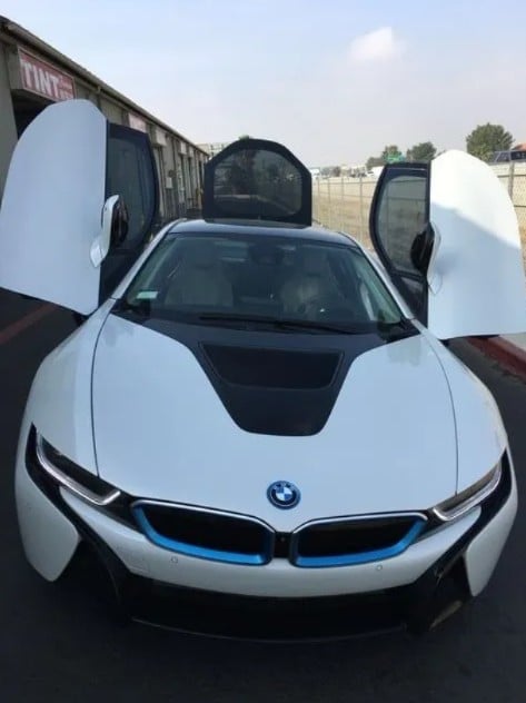 White BMW i8 hybrid sports car with open butterfly doors, blue accents, photographed front-on in a parking lot