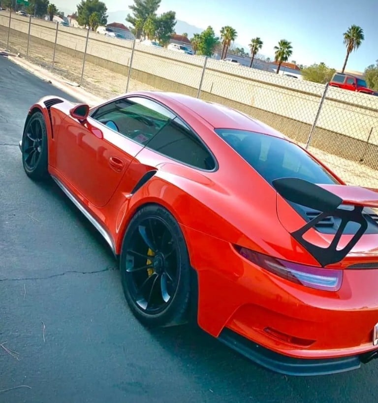 Red sports car, possibly a Porsche, parked in a lot with palm trees and buildings visible in the background