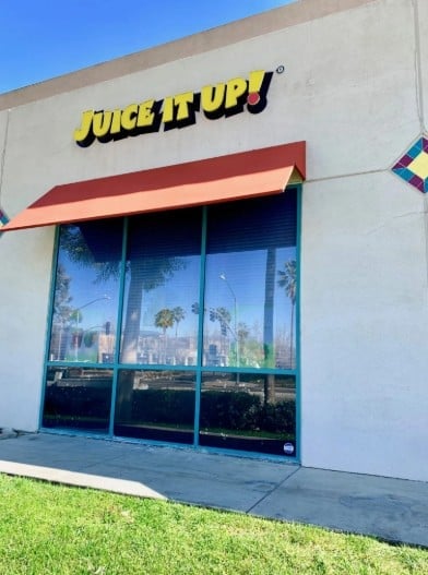 Juice It Up storefront with yellow logo sign, red awning, and large glass doors reflecting palm trees and city skyline