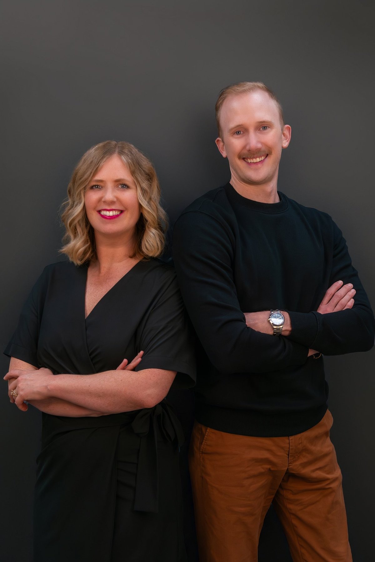 Professional headshot of a smiling woman with blonde wavy hair and a man with reddish hair, both wearing dark tops against a gray background