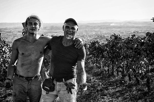Two men smiling with arms around each other in a vineyard with a cityscape in the background, black and white photo