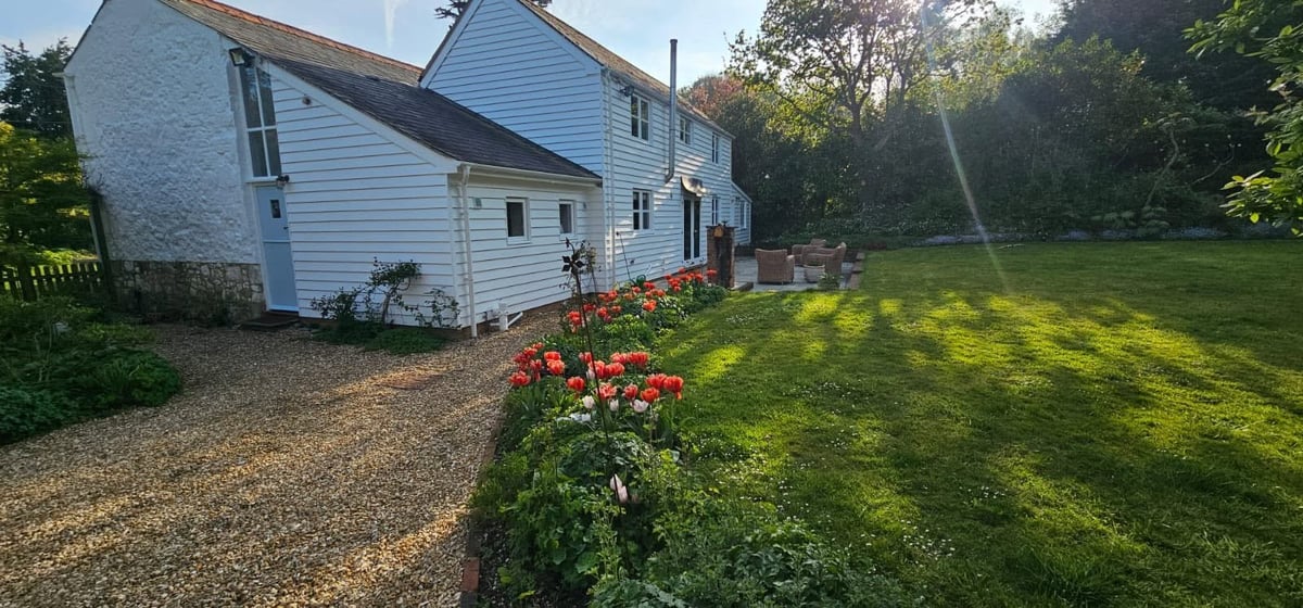 White farmhouse with blue shutters, gravel driveway, red flowers, and expansive green lawn with trees in background