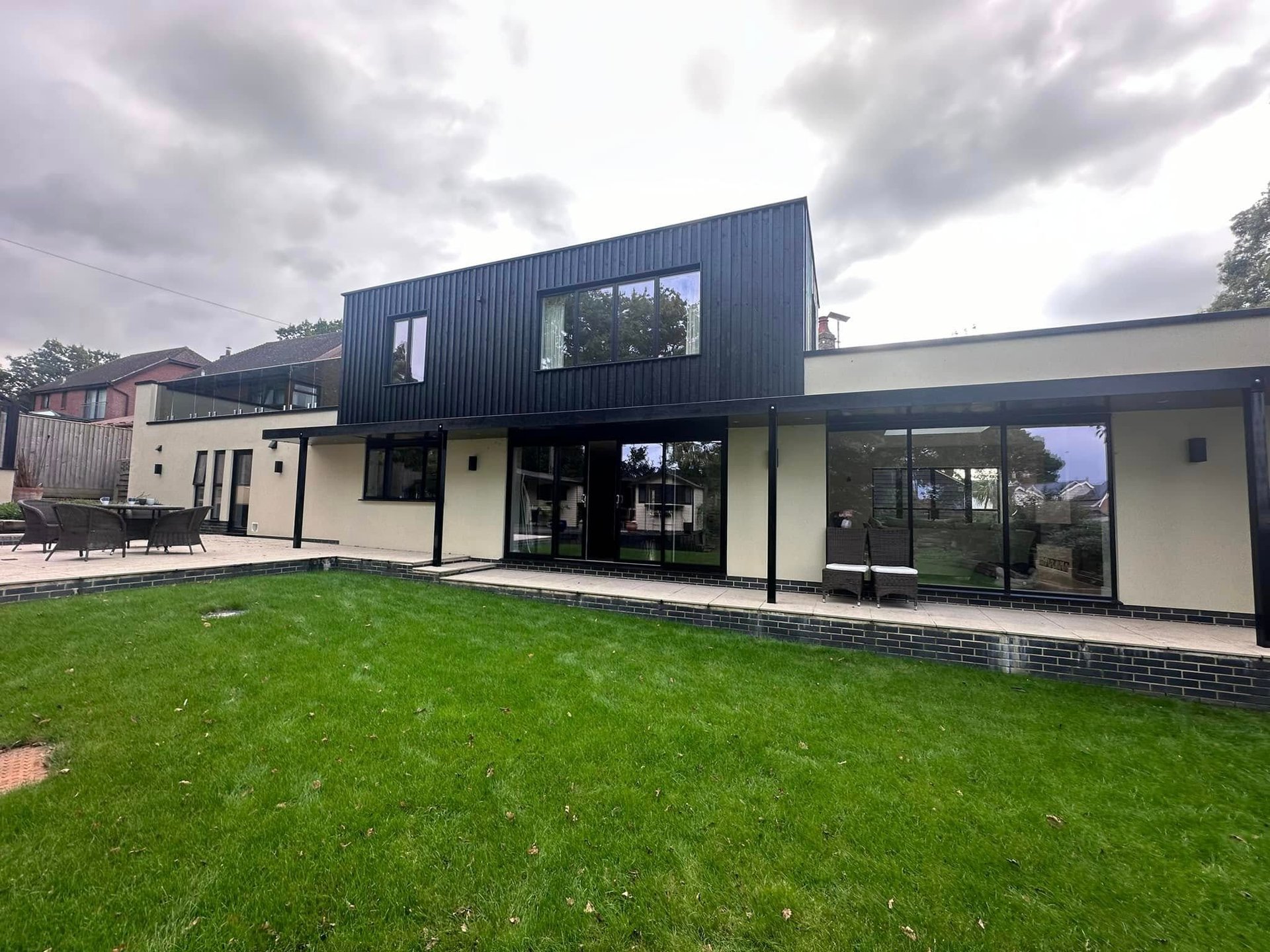 Modern black and cream two-story house with large windows, green lawn, and cloudy sky