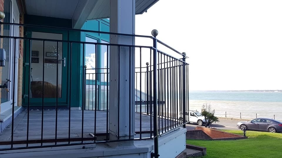 Modern beachfront house with black metal railing balcony overlooking ocean, parked cars, and green lawn