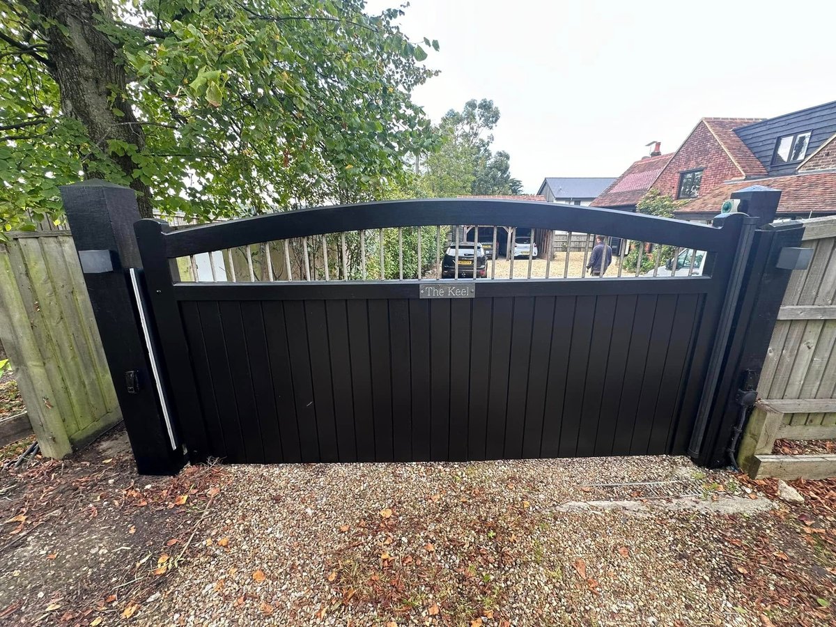Black metal driveway gate with bars and lattice detailing on a gravel entrance with residential houses visible in background