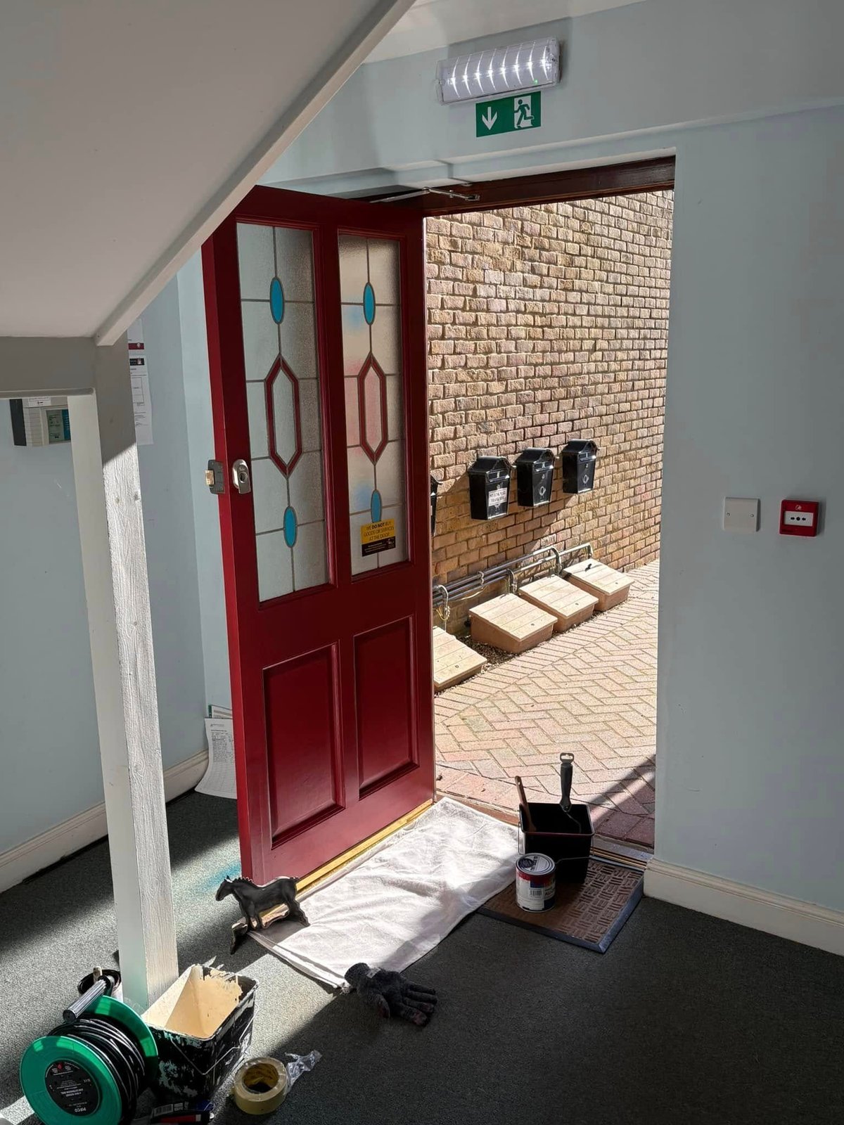 Red entrance door with decorative glass panels, open to a brick exterior area with emergency exit sign and equipment stored outside