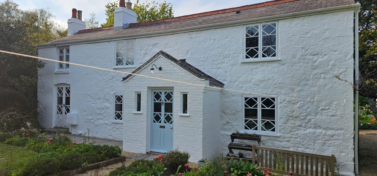 White painted cottage with pale blue door, multiple windows, two chimneys, and wooden bench in front garden