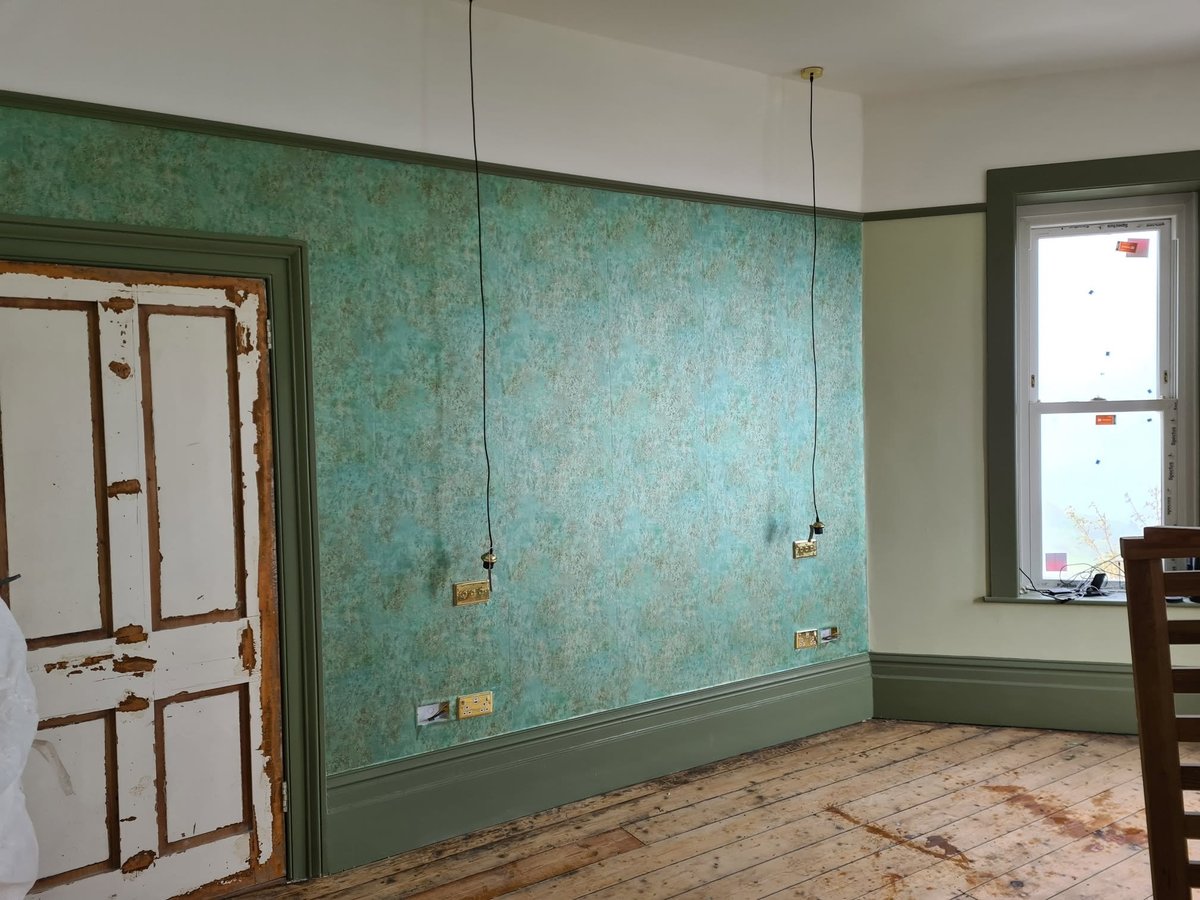 Empty room with turquoise patterned wallpaper, wooden door, window, pendant light fixtures, and wooden floorboards