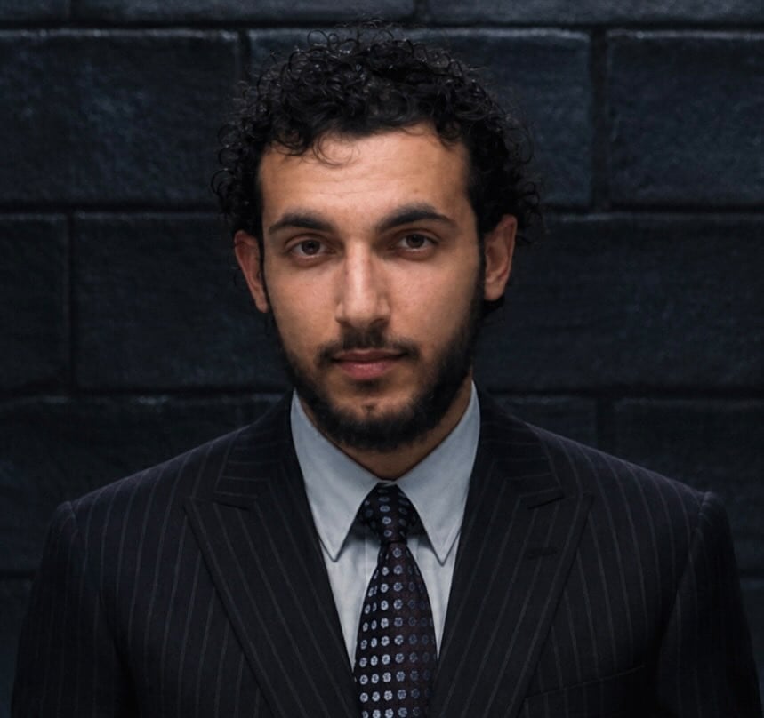 Man in dark pinstriped suit and patterned tie standing against a dark tiled background, looking directly at camera