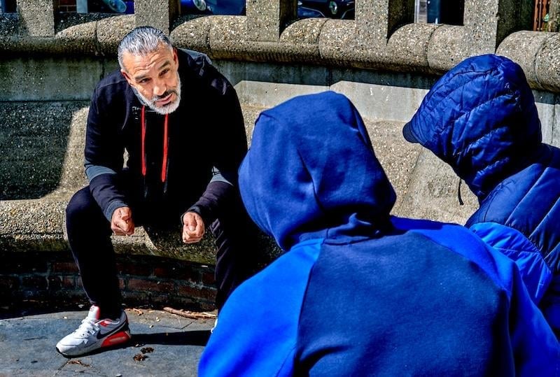 Older man in black jacket crouching while speaking with two people wearing bright blue hoodies