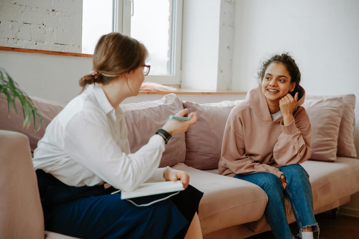 Two women having a conversation on a beige couch in a bright living room with large windows