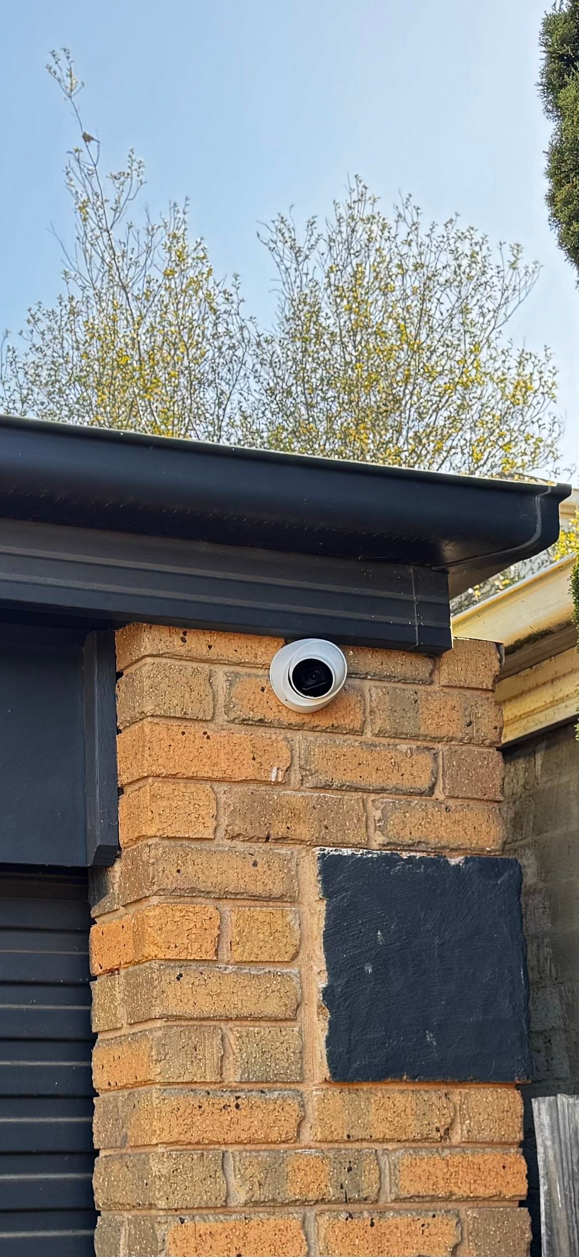 Security camera mounted on brick wall corner of dark roof with budding trees in background at a Melbourne property