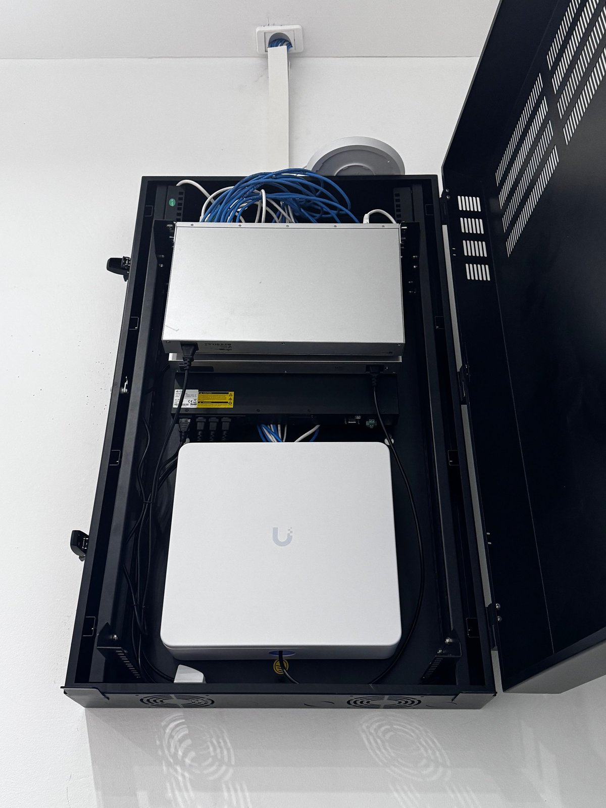 Commercial network server cabinet with white networking equipment, blue ethernet cables, and cooling fan mounted on a white wall