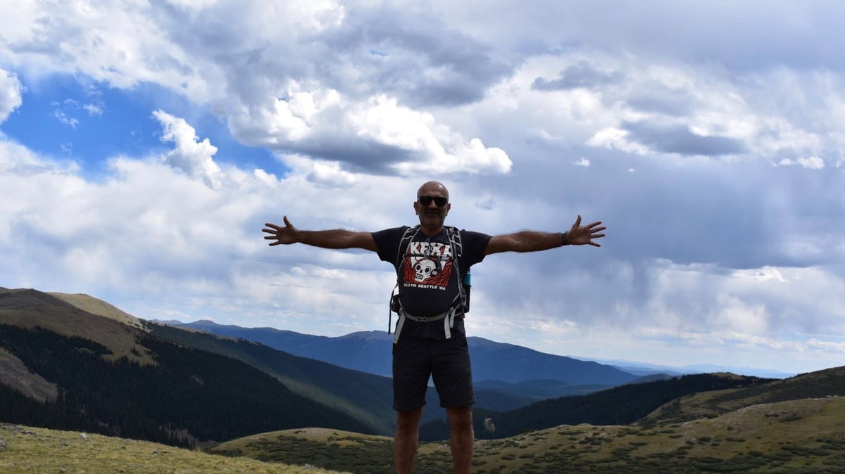 Person standing with arms outstretched on a grassy hillside overlooking mountains under a cloudy sky