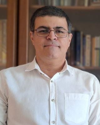 Middle-aged man with short dark hair and glasses wearing a white button-up shirt, photographed indoors with bookshelves in the background