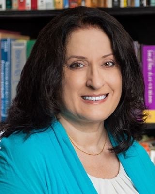Professional headshot of a woman with long dark hair wearing a turquoise cardigan and white top, smiling at camera with bookshelves in background