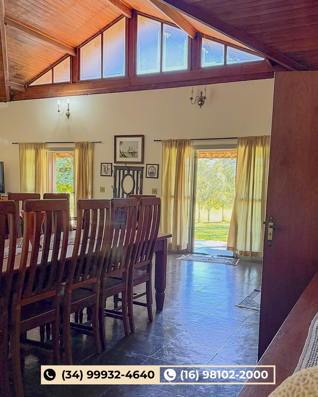 Sala de jantar ampla com vigas de madeira, janelas no teto, cadeiras escuras, cortinas amarelas e piso de pedra com acesso ao pátio