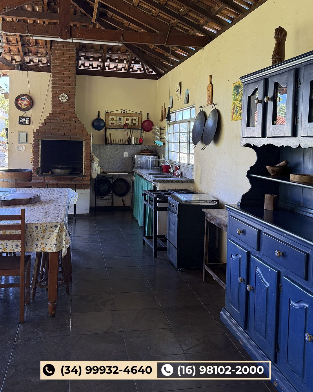 Cozinha rústica com armários azuis, lareira de tijolinho, panelas suspensas e mobília de madeira em ambiente rural