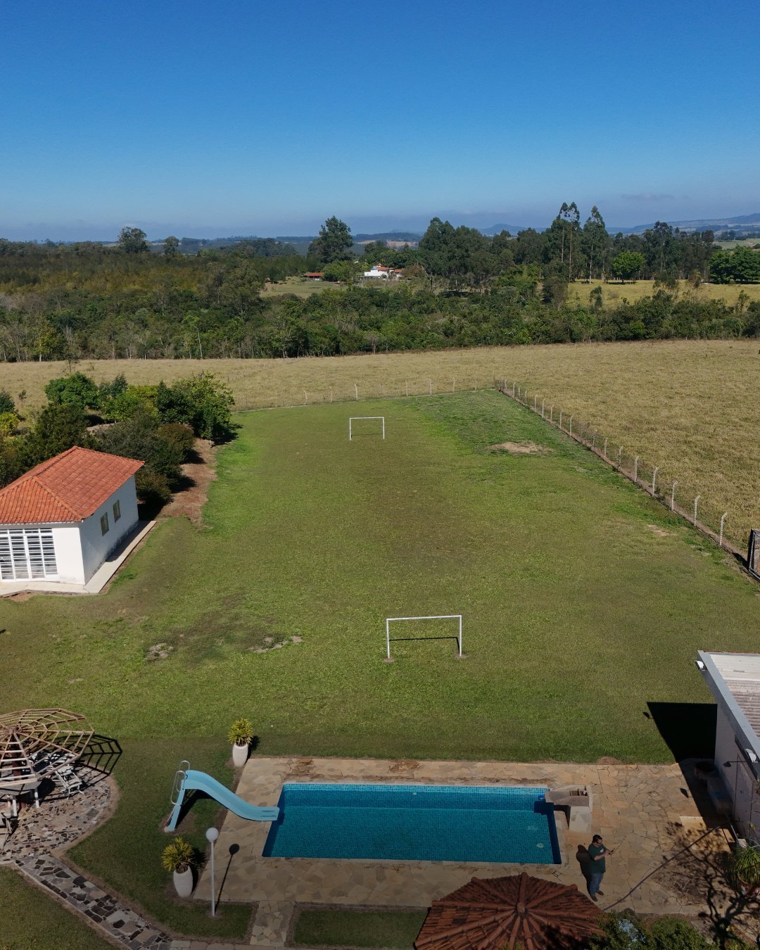 Vista aérea de propriedade com piscina, traves de campo de futebol em gramado e construção branca de telhado vermelho