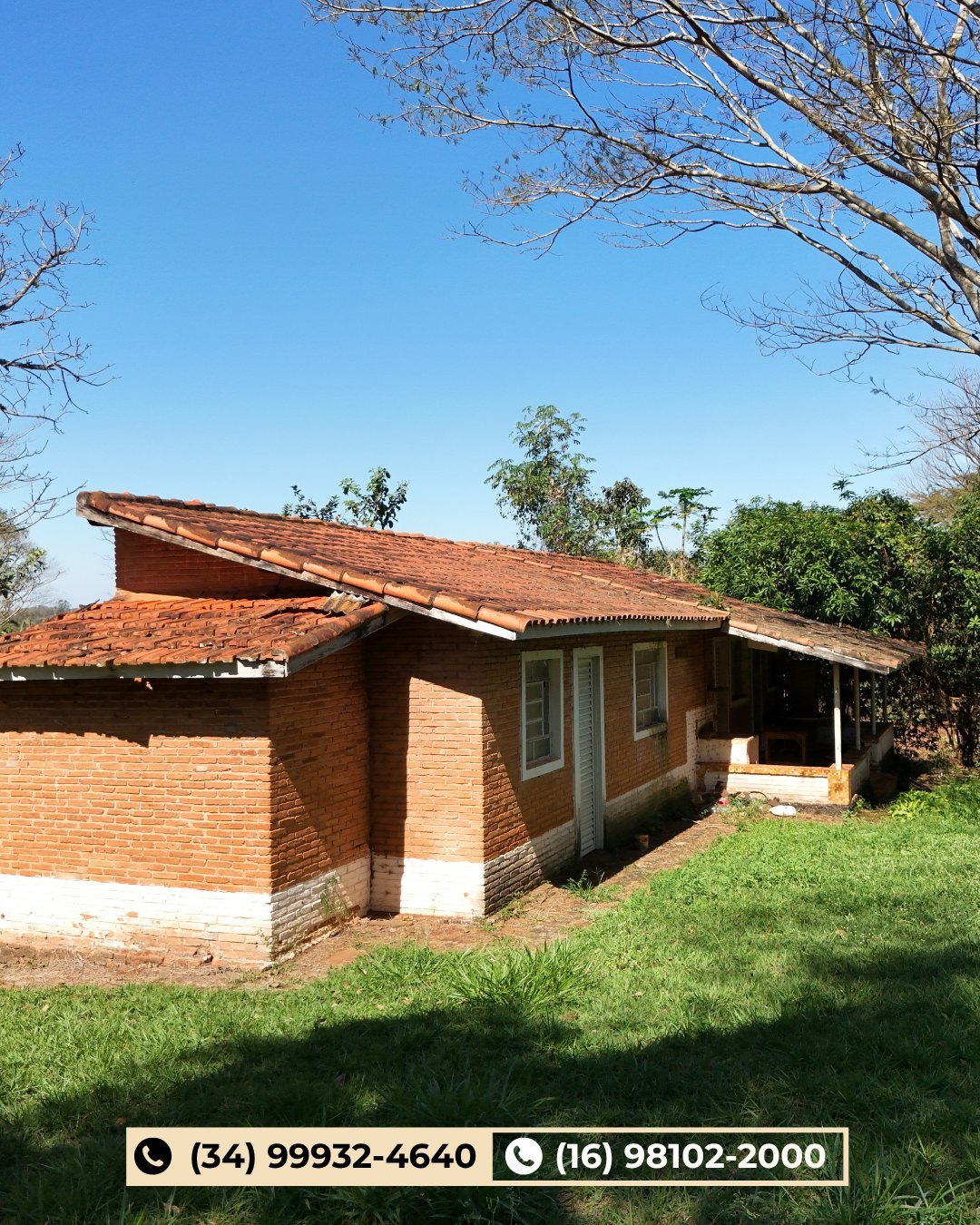 Casa rústica de tijolinho com telhado de barro, cercada por gramado verde e árvores sob céu azul