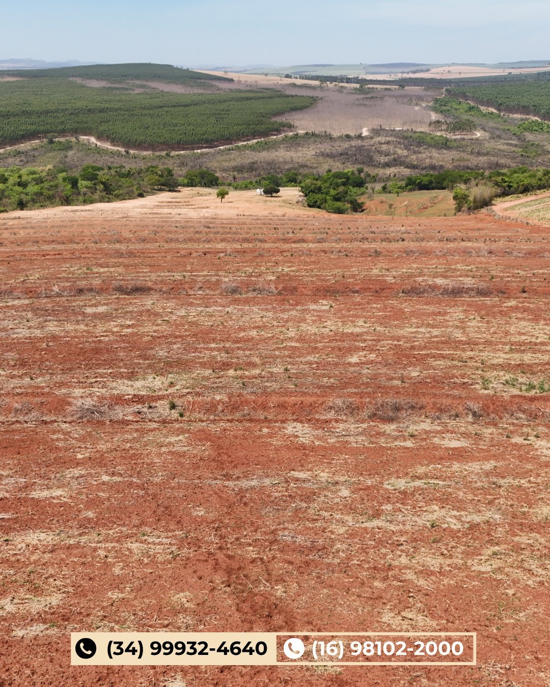 Vista aérea de área rural com solo avermelhado em primeiro plano, campos agrícolas verdes e árvores ao fundo sob céu limpo