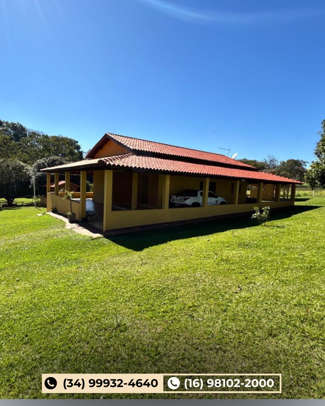 Casa sede amarela com telhado vermelho sobre gramado verde cercada por árvores sob céu azul