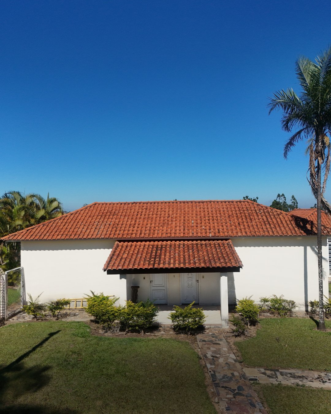 Casa branca com telhado de barro e varanda coberta cercada por palmeiras e gramado sob céu azul