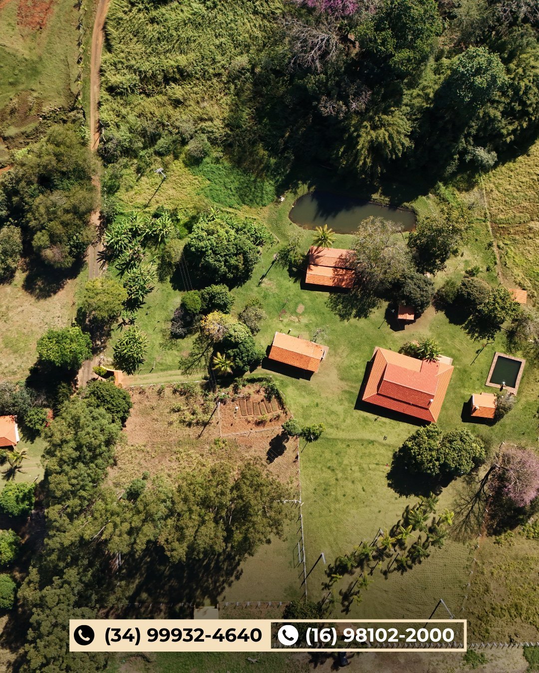 Vista aérea do sítio com construções de telhado vermelho, pastagem verde, árvores e lagos cercados por floresta