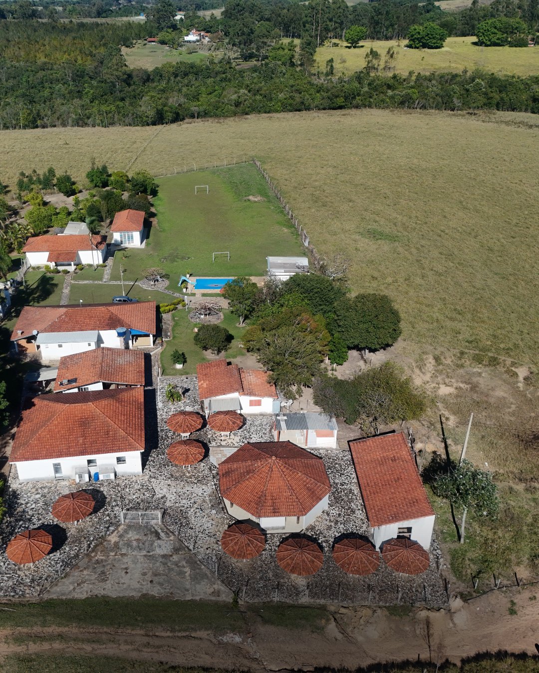 Vista aérea de propriedade rural com construções brancas, telhados coloniais, piscina e campo de futebol cercados por lavouras
