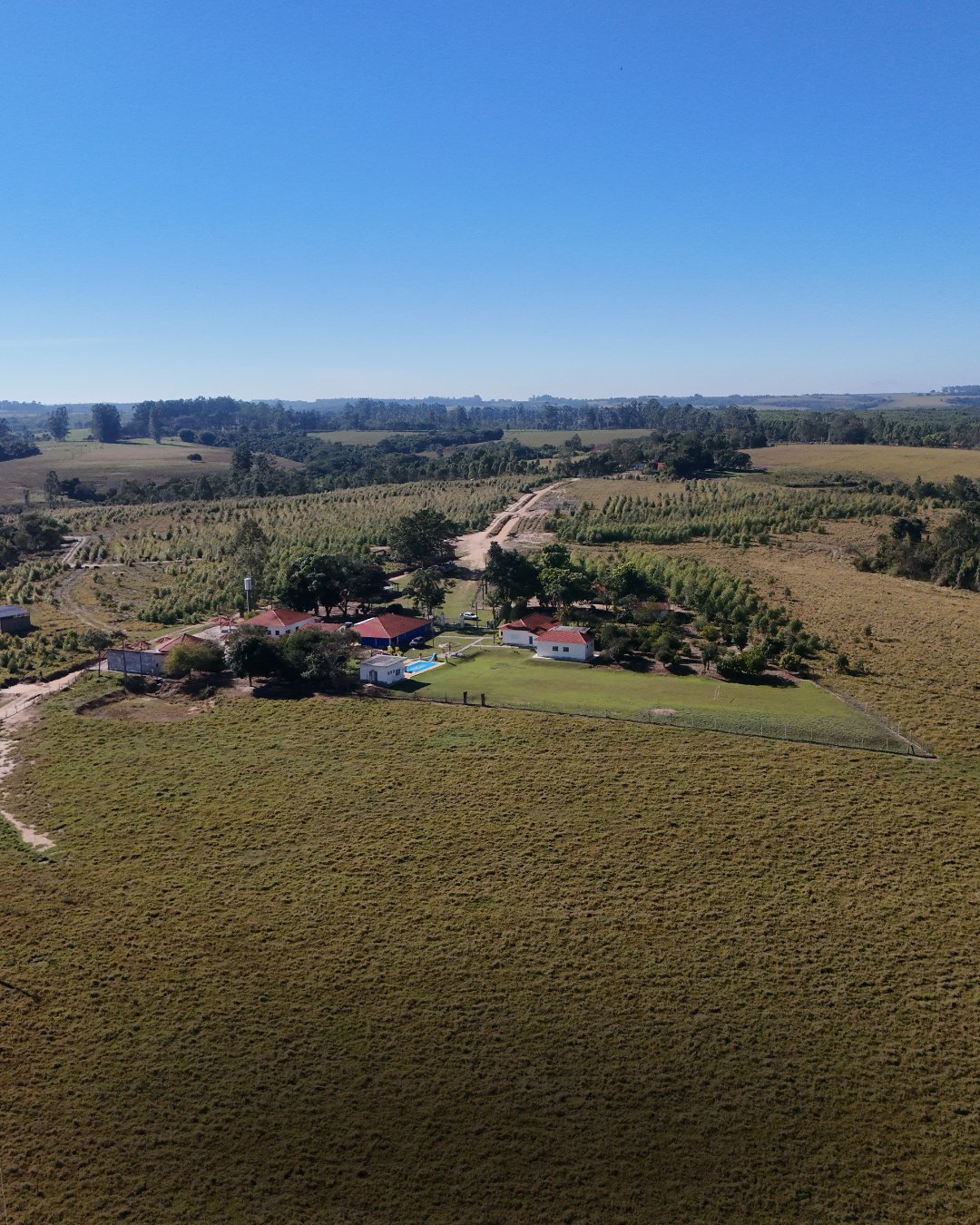 Vista aérea de sede rural com telhados vermelhos cercada por campos verdes, pomares e bosques sob céu azul