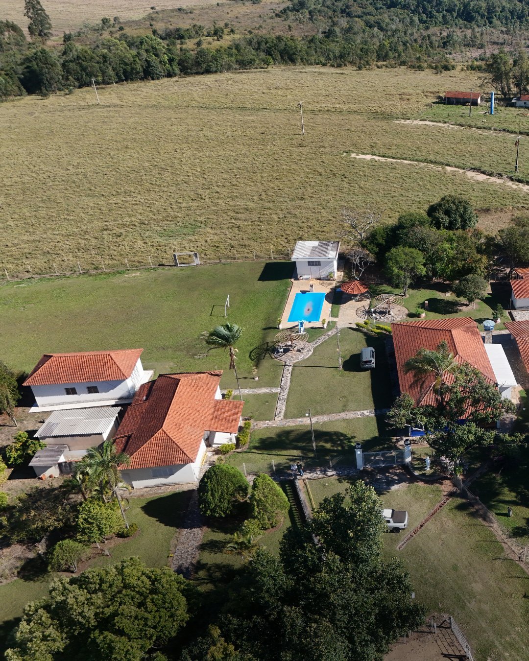 Vista aérea de propriedade rural com casas de telhado vermelho, piscina azul, gramado e áreas de cultivo ao redor