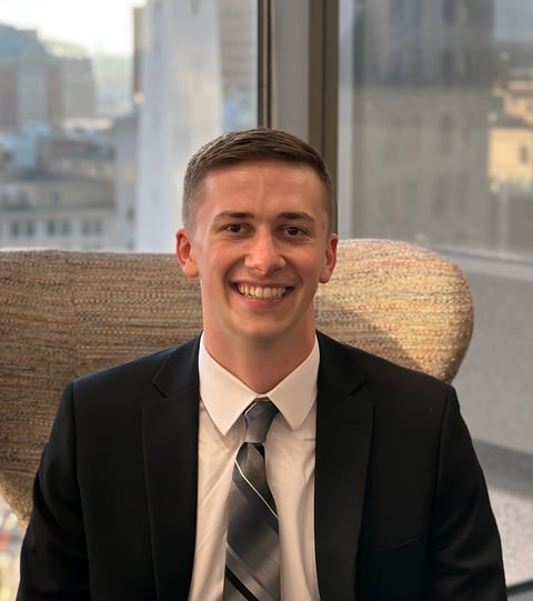 Smiling man in business suit and tie sitting in office chair with city skyline visible through windows behind him