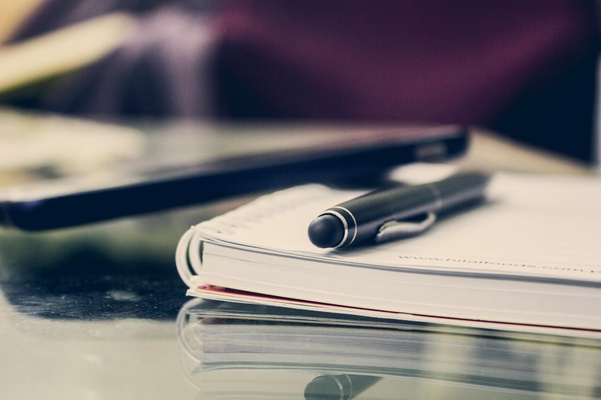 A pen resting on an open notebook with a smartphone blurred in the background on a desk