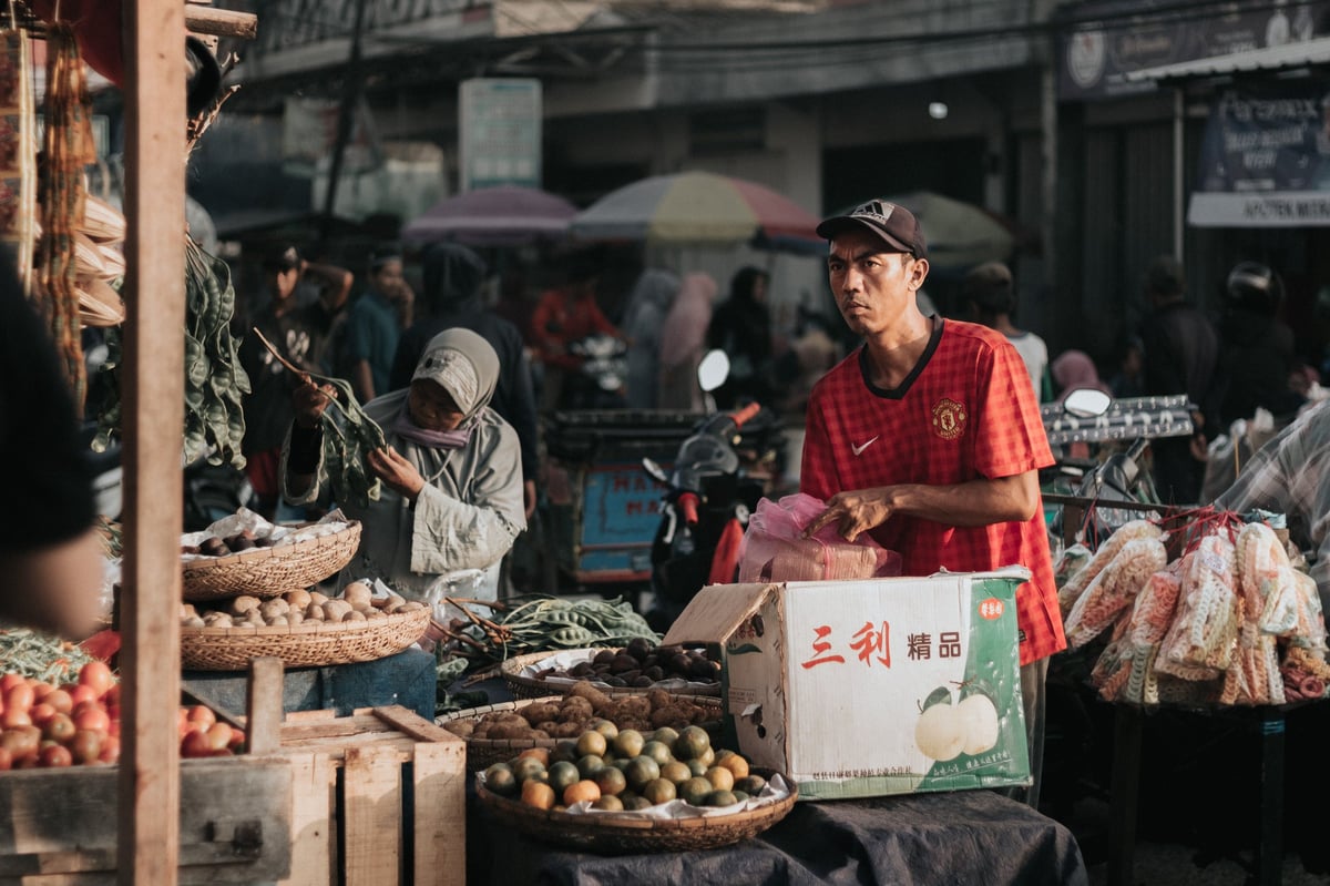 Vendor in red shirt standing at outdoor market stall with fresh vegetables and produce displayed.