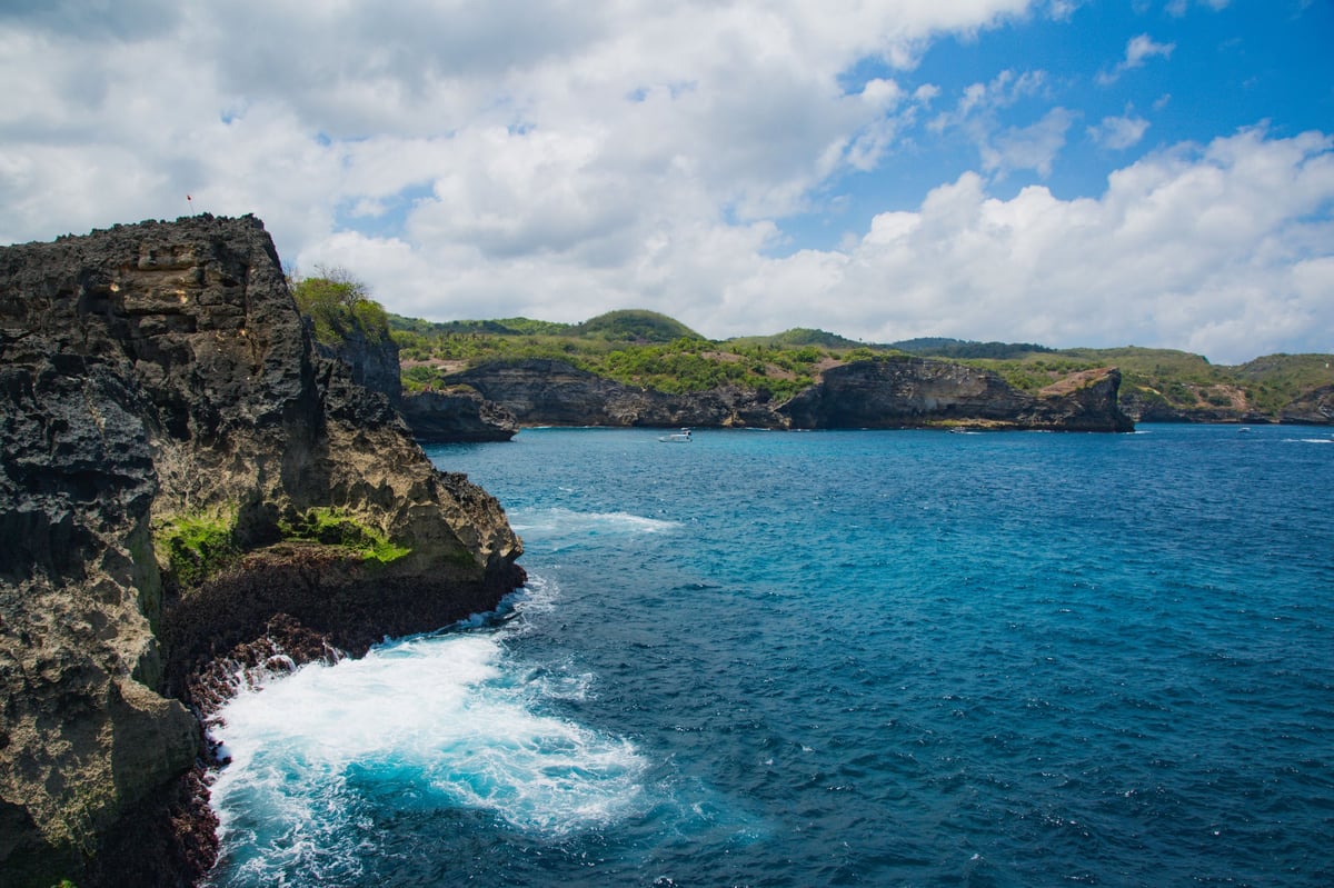Dramatic rocky cliffs and turquoise ocean with white waves under a blue sky with clouds.