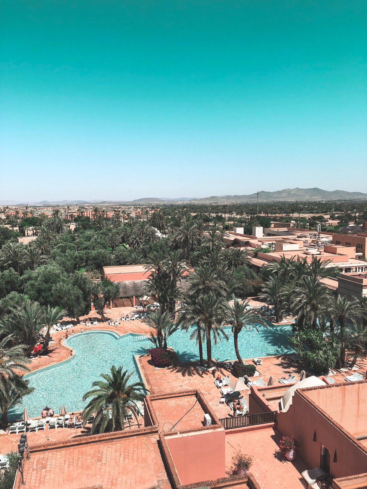 Aerial view of a resort pool surrounded by palm trees with terracotta buildings and a desert city landscape under blue sky