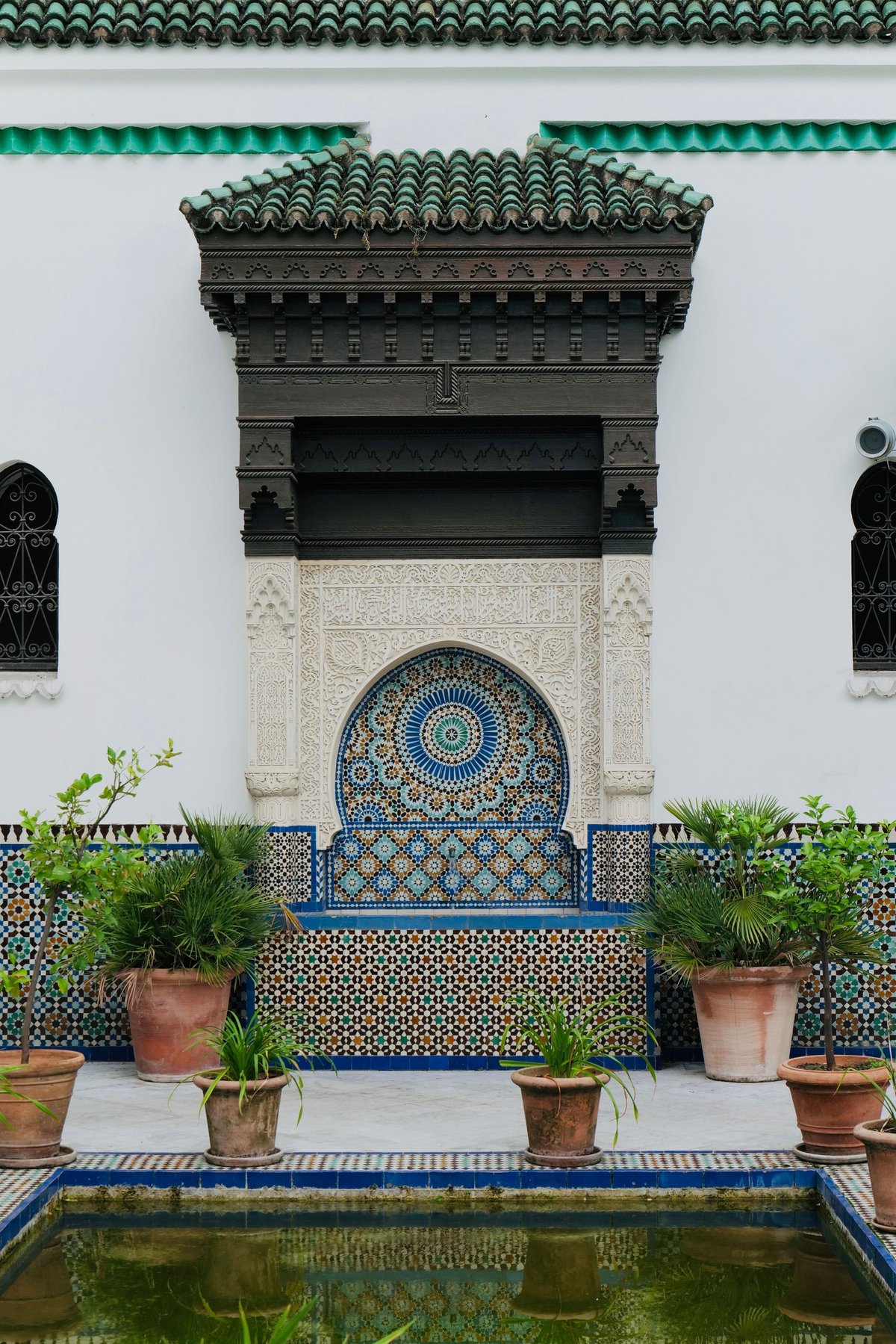 Moroccan courtyard with white walls, ornate green-tiled roof, decorative blue geometric fountain niche, terracotta pots, and reflective water pool