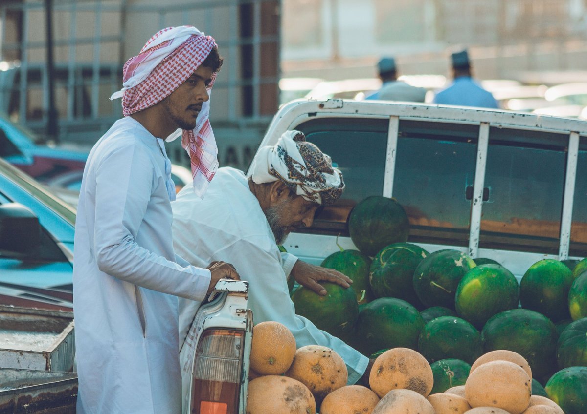 Two men in traditional Middle Eastern dress examining produce at a waterside market stall with melons and other fruits