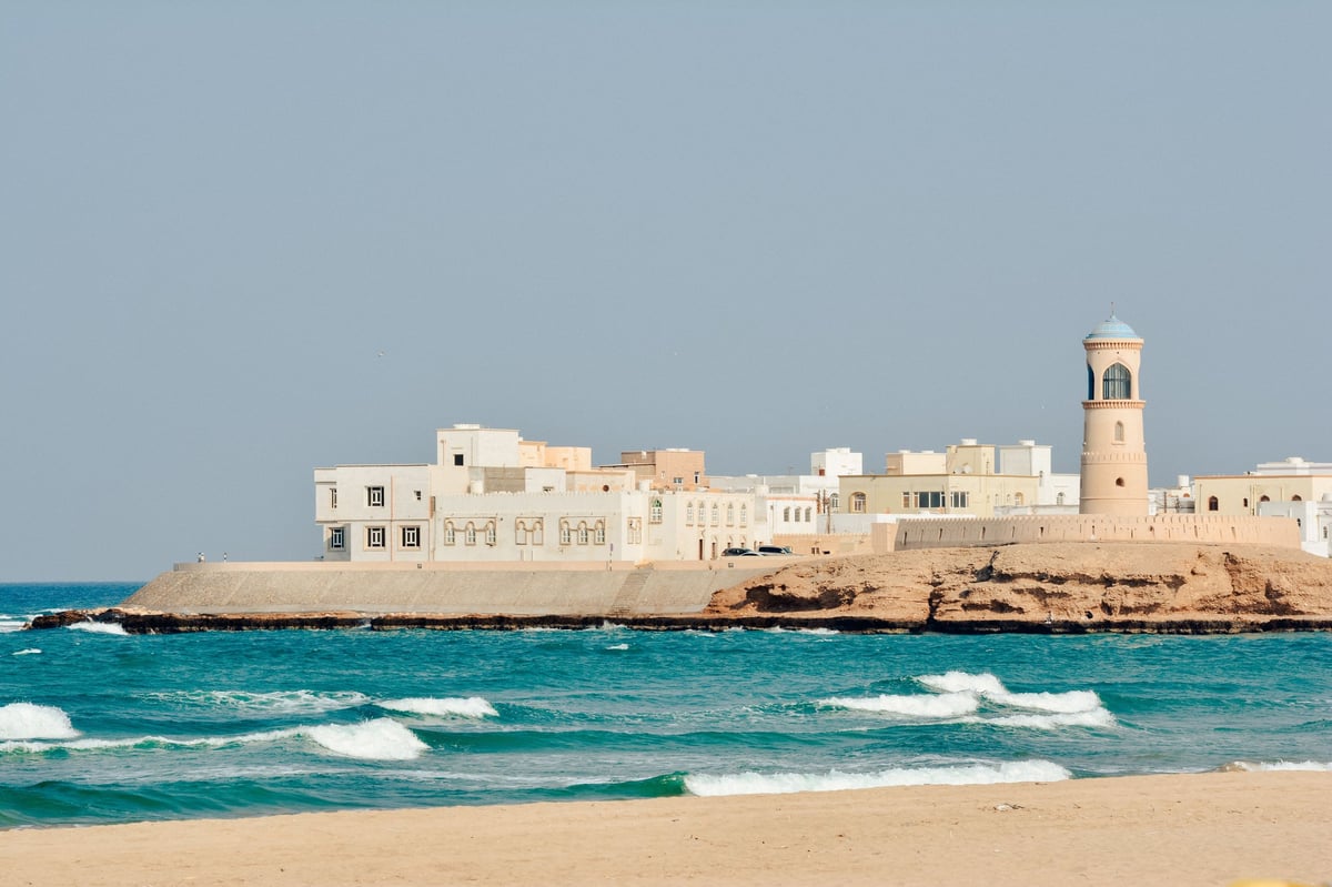 Walled coastal fortress with lighthouse overlooking turquoise sea and sandy beach with white-capped waves